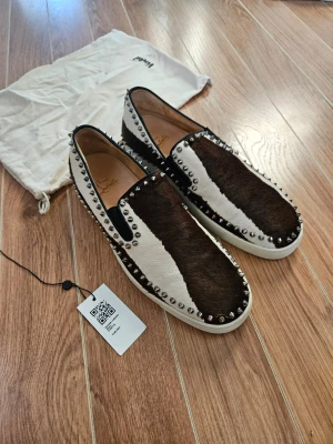 Christian Louboutin Rolle Boat Horse Hair Slip Ons - Unika slip-on sneakers från Christian Louboutin med svart och vit häst päls, dekorerade med silverfärgade nitar runt hela skon. Skorna har en vit sula och den klassiska röda undersidan. Perfekt för dig som vill sticka ut med din stil. I stort sett 1 of 1 modell som inte längre tillverkas eller säljs. Saknar en nit vänster bak, syns på sista bilden men inte när man har på sig dem.