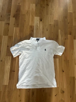 Vit kortärmad pikétröja - Klassisk vit pikétröja med korta ärmar och krage. Tröjan har knappar vid halsen och en liten broderad logga på bröstet. Perfekt till jeans eller shorts för en clean och stilren look. Från Polo ralph lauren.