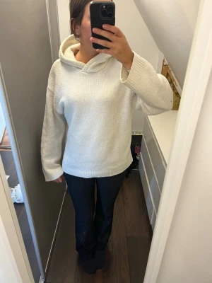 Vit stickad hoodie med huva - Mysig vit stickad hoodie med avslappnad passform och stor huva. Tröjan har långa ärmar och ribbade muddar vid ärmslut och nederkant. Perfekt för dig som gillar en enkel och clean stil. Materialet ser ut att vara mjukt och lite fluffigt.
