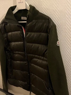 Grön cardigan från Moncler - Snygg grön cardigan från Moncler med stickade ärmar och quiltad front i blankt material. Jackan har hög krage, dragkedja framtill och Moncler-logga på ärmen. Perfekt för kalla dagar och ger en cool streetwear-look.