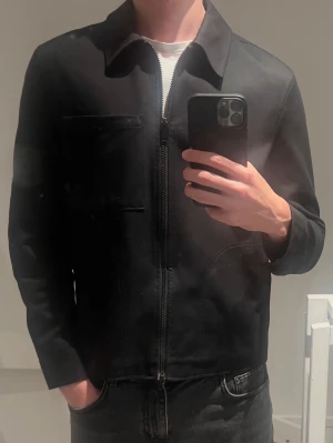 Svart jacka från Zara - Stilren svart overshirt från Zara med rak passform och dragkedja framtill. Overshirten har en bröstficka och klassisk krage. Tillverkad i ett slätt tyg som ger en clean look. Perfekt att bära över en t-shirt för en enkel och snygg stil.