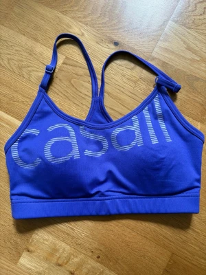 Blå sport-bh från Casall - Snygg lila sport-bh från Casall 💜 med justerbara axelband och bred resår nedtill. Bh:n har ett stort tryck med Casall-loggan framtill och är tillverkad i ett stretchigt funktionsmaterial som passar perfekt till träning. Vadderade kupor i storlek xs 