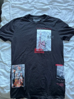 Svart t-shirt Supply & Demand NYC - Svart t-shirt från Supply & Demand med tryck av Frihetsgudinnan och texten 'NYC' samt 'Supply Demand' i rött framtill. T-shirten har korta ärmar och flera grafiska detaljer i vitt, rött och grått. Perfekt för dig som gillar streetwear och coola prints.