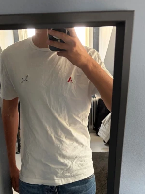 Vit t-shirt med röd A-detalj - Säljer en vit t-shirt med korta ärmar och rund hals. På bröstet finns en liten röd broderad bokstav A samt en silverfärgad detalj bredvid. T-shirten har en clean och enkel look som passar till jeans eller shorts.