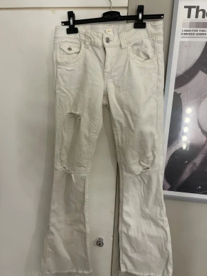 Vita bootcut jeans med slitningar - Säljer ett par vita bootcut jeans med slitna detaljer på benen och råa kanter nertill. Jeansen har fem fickor, knappgylf och är tillverkade i ett mjukt bomullstyg. Perfekta för en avslappnad och trendig look.