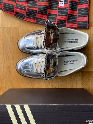 Adidas Wales Bonner silvriga sneakers - Unika sneakers från Adidas x Wales Bonner med metallic-silver ovandel, vita snören och bruna detaljer på plösen. Skorna har låg profil och klassisk retrostil med kontrasterande sömmar och Wales Bonner branding på innersulan. Perfekta för dig som vill sticka ut.
