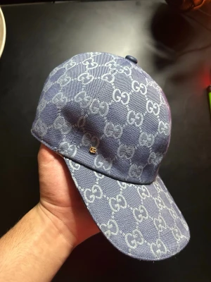Blå GG-monogram keps från Gucci - Säljer en blå keps från Gucci med det klassiska GG-monogrammet över hela kepsen. Kepsen är i denimliknande material och har justerbar rem bak. Insidan är ljus och har Gucci-taggar. Perfekt för dig som vill ha en stilren och lyxig accessoar.