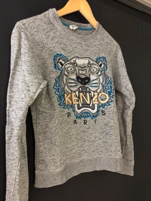 Grå sweatshirt med tiger från Kenzo - Grå sweatshirt från Kenzo med ikonisk broderad tiger och texten 'KENZO PARIS' på bröstet. Tröjan har rund hals, långa ärmar och ribbade muddar. Perfekt för dig som gillar streetwear och vill sticka ut med en cool detalj framtill.