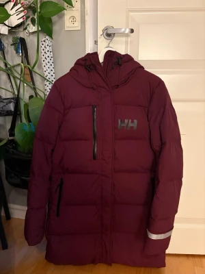 Vinröd dunjacka från Helly Hansen - Aldrig andvänd vinröd dunjacka från Helly Hansen med huva och dragkedjor. Jackan har en längre modell, reflekterande detalj på ärmen och HH-logga på bröstet. 