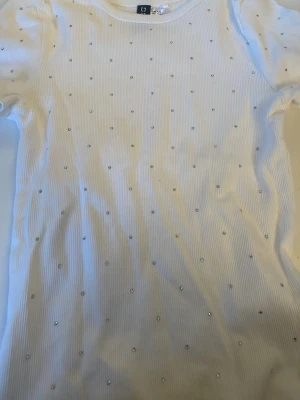 Vit ribbad topp med pärlor från HM - Säljer en vit ribbad topp från HM, dekorerad med små pärlor över hela framsidan. Toppen har korta ärmar och en rund halsringning. Perfekt för dig som gillar detaljer och en clean look.