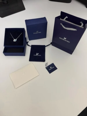 Swarovski halsband med kristallhjärta - Säljer ett elegant halsband från Swarovski med ett gnistrande hjärthänge i klar kristall. Kedjan är silverfärgad och halsbandet kommer med originalask, påse och certifikat. Perfekt accessoar för dig som gillar stilrena smycken med lite extra bling.