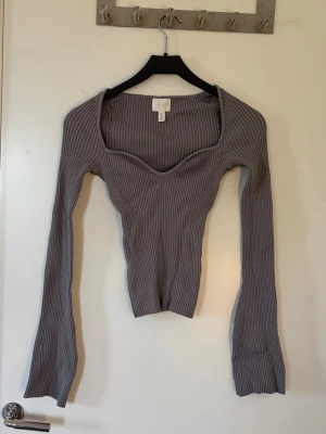 Grå ribbad långärmad topp - Säljer en grå ribbad topp med lång ärm och snygg, formad urringning. Toppen är figurnära och har en modern, croppad passform som passar perfekt till jeans eller kjol. Materialet är stretchigt och bekvämt.