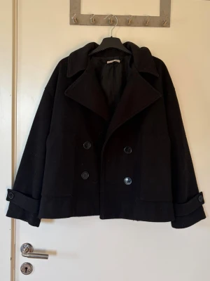 Svart dubbelknäppt kappa - Svart skepparkavaj med dubbelknäppning, bred krage och stora fickor framtill. Jackan har uppvikta ärmslut med knappdetalj och är tillverkad i ull för en klassisk look. Passformen är loose och modellen är något croppad.