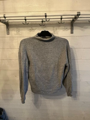 Grå stickad tröja från Gina Tricot - Mysig grå stickad tröja från Gina Tricot med hög hals och långa ärmar. Perfekt för lager-på-lager och enkel att matcha med jeans eller kjol. Tröjan har en klassisk rak passform och är superbekväm.