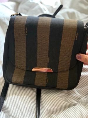 Randig handväska från Fendi - Snygg handväska från Fendi med breda svarta och bruna ränder. Väskan har ett guldfärgat metallspänne med Fendi-logga och justerbar axelrem. Tillverkad i slitstarkt tyg med klassisk design och subtila Fendi-loggor i mönstret. ÄKTA, inget kvitto då den är köpt för många år sen
