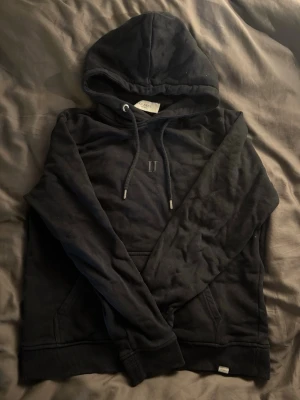 Svart hoodie från Les deux med ficka - En svart hoodie med huva och dragsko, samt en klassisk magficka framtill. Tröjan har ribbade muddar vid ärmslut och nederkant. Materialet ser ut att vara mjukt och bekvämt, perfekt för chill dagar. Diskret broderad detalj på bröstet.