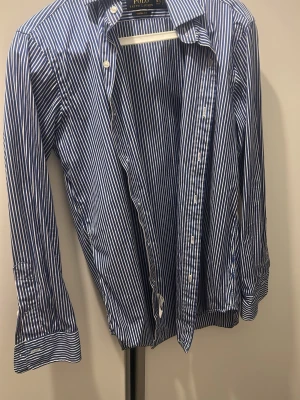 Blåvit randig skjorta från Polo Ralph Lauren - Klassisk blåvit randig skjorta från Polo Ralph Lauren med lång ärm och knappar framtill. Skjortan har en tidlös design med smal krage och manschetter. Perfekt till jeans eller chinos för en clean och stilren look. Storlek M