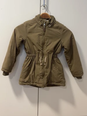 Beige parkasjacka med snörning - Snygg beige parkasjacka med dragkedja och snörning i midjan. Jackan har två fickor framtill och är fodrad med mjuk fleece upptill och quiltat tyg nedtill. Perfekt för kyligare dagar och har en klassisk, tidlös look.