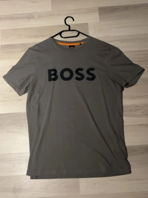 Hugo Boss T-shirt - Snygg grå t-shirt från BOSS med stort svart logotryck över bröstet. Klassisk rund halsringning och korta ärmar. Tillverkad i mjuk bomull som känns skön mot huden. Perfekt för dig som gillar stilrena och enkla plagg med tydlig branding.