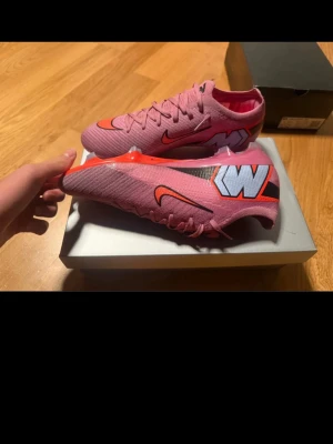 Rosa Nike Mercurial fotbollsskor - Säljer ett par rosa Nike Mercurial fotbollsskor med orange detaljer och svart-vit grafik på sidan. Skorna har en låg profil, snörning och en yttersula med dobbar för bra grepp på planen. Tillverkade i syntetmaterial för lätt känsla och snabbhet.