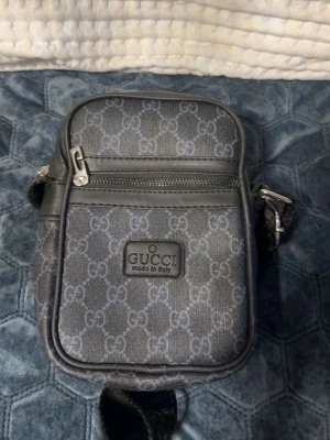 Svart axelväska från Gucci - Snygg svart axelväska från Gucci med klassiskt GG-mönster i grått. Väskan har en justerbar axelrem, dragkedja framtill och detaljer i svart läder. Perfekt storlek för det viktigaste och en stilren look med tydlig logotyp på framsidan.