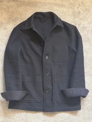 Mörkblå overshirt - Stilren mörkblå overshirt med tre utanpåliggande fickor och knäppning framtill. Overshirten har klassisk krage och uppvikta ärmslut. Materialet ser ut att vara tjockt och strukturerat, perfekt för lager på lager. Enkel och clean design som passar till det mesta.