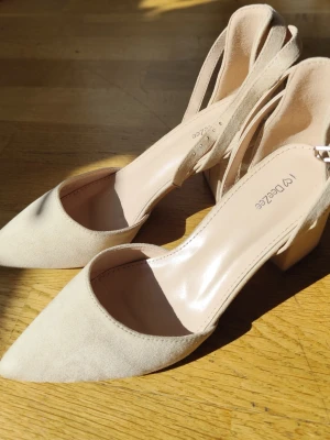 Beige pink pumps med blockklack - Snygga beige pink pumps från Dezzee med spetsig tå och bred blockklack. Skorna har öppen häl med justerbart spänne och är tillverkade i mockaimitation. Perfekta för dig som vill ha både stil och komfort.