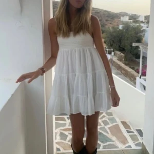 Vit klänning Zara - Supersöt vit kortklänning med tunna axelband och volanger i flera lager. Klänningen har en luftig och flowy passform som ger en chill vibe, perfekt för varma dagar. Materialet känns lätt och fräscht, och den är ärmlös för en somrig look.