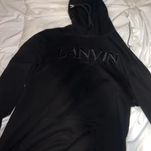 Svart Lanvin hoodie med broderad logga - Säljer en svart hoodie från Lanvin med broderad logga på bröstet. Tröjan har huva med snörning och en stor magficka framtill. Perfekt för dig som gillar stilrena och exklusiva plagg med streetkänsla.