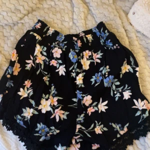 Svarta blommiga shorts med spetskant - Säljer ett par svarta shorts med blommigt mönster i ljusrosa, blått och gult. De har en bred, smockad midja och är dekorerade med en fin spetskant längst ner. Luftiga och sköna, perfekta för varma dagar.
