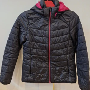 Svart pufferjacka från Everest 146/152 - Svart pufferjacka från Everest med rosa foder och dragkedja. Jackan har huva, quiltad design och två fickor med dragkedja. Perfekt för kyliga dagar och har en sportig vibe. Materialet är syntet och jackan är lätt och smidig att bära.