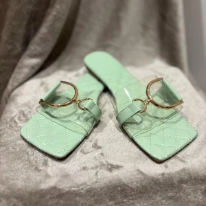 Mintgröna sandaler med gulddetalj - Snygga mintgröna sandaler med quiltad sula och fyrkantig tå. De har transparent rem över foten och en stor guldfärgad metallspänne som detalj. Perfekta för att lyfta din sommarlook med en trendig touch.