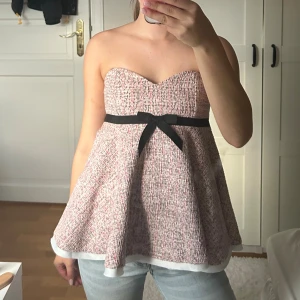 Rosa tubtopp med svart rosett - Säljer en söt tubtopp i ljusrosa och vit tweed med svart band och rosett under bysten. Toppen har en hjärtformad urringning och är utsvängd nertill med vit kant. Axelbanden är avtagbara så du kan bära den både axelbandslös och med band.