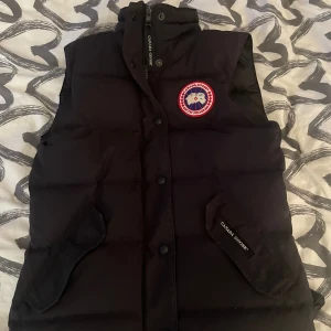 Svart dunväst från Canada Goose XS - Svart dunväst från Canada Goose med klassisk Arctic Program-logga på bröstet. Västen har hög krage, tryckknappar och dragkedja framtill samt två fickor med lock. Materialet är polyester och dun, perfekt för kyliga dagar. Snygg och tidlös design med quiltad yta.