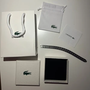 Silverfärgat armband från Lacoste - Stilrent armband i silverfärgad metall från Lacoste med klassisk krokodillogga på låset. Länkarna har ett snyggt mönster som ger extra edge. Levereras med originalförpackning, tygpåse och äkthetsbevis. Perfekt accessoar för dig som gillar sportig och modern stil.