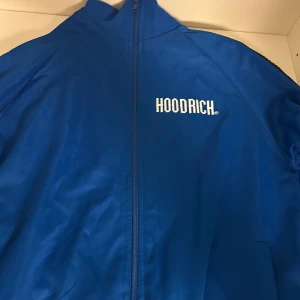 Blå Hoodrich tröja - Snygg blå jacka från Hoodrich med dragkedja och tryckt logotyp på bröstet. Jackan har svarta detaljer längs ärmarna med upprepade Hoodrich-logotyper. Perfekt för en sportig look. Den är nästan som ny då jag har använt ett par gånger bara.