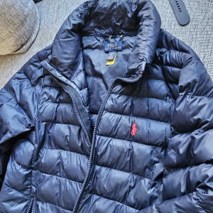 Mörkblå pufferjacka Polo Ralph Lauren - Snygg mörkblå pufferjacka från Polo Ralph Lauren med klassisk röd logga på bröstet. Jackan har quiltad design, hög krage och dragkedja framtill. Lättviktsdun och passar kvinna xs eller s och barn från 164/170. Storlek XL/16 år i Ralph lauren storlek. Väldigt fint skick hittar inga fel!