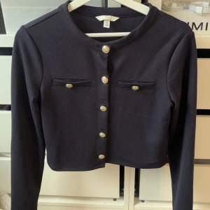 Mörkblå croppad kofta från H&M - Snygg mörkblå croppad kofta från H&M med guldfärgade knappar framtill och två dekorativa fickor på bröstet. Koftan har lång ärm och rund halsringning, perfekt för att ge din outfit en chic touch.