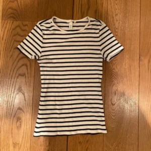 Randig vit och svart t-shirt - Snygg vit t-shirt med svarta horisontella ränder och rund hals. Modellen är klassisk med korta ärmar och en slim passform som sitter nära kroppen. Perfekt för dig som gillar enkel och stilren design.PRISET GÅR ATT DISSKUTERA 