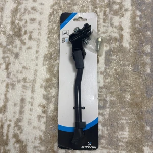 B'TWIN cykelstöd 16-20 tum - Stabilt cykelstöd från B'TWIN, passar cyklar med hjulstorlek 16-20 tum. Produkten är ny i originalförpackning och inkluderar monteringsskruvar. Perfekt för att enkelt parkera din cykel. Inga synliga defekter eller slitage.