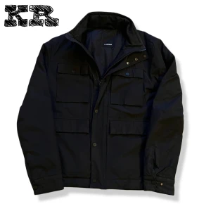 j.lindeberg feildjacket - Svart vindjacka från J.Lindeberg med fyra stora fickor framtill, dragkedja och tryckknappar. Jackan har en klassisk krage och diskret JL-logga på ärmen. Materialet är slitstark polyester och passformen är normal. Perfekt för dig som gillar stilren och funktionell design.