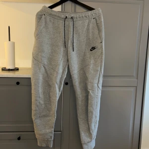 Grå mjukisbyxor från Nike, storlek M - Snygga grå mjukisbyxor från Nike med svarta detaljer och dragsko i midjan. Byxorna har ribbade muddar vid bensluten, praktiska fickor och en liten Nike-logga på ena benet. Perfekta för chill eller träning.