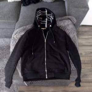 Svart Burberry hoodie med dragkedja - Svart hoodie från Burberry med klassiskt rutigt foder i huvan och insidan. Tröjan har dragkedja framtill, snörning i huvan och broderad logga på bröstet. Tillverkad i mjuk bomull, perfekt för en chill och snygg look.