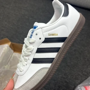 Adidas Samba vita och svarta sneakers - Adidas Samba sneakers i vitt skinn med svarta detaljer och klassiska tre ränder på sidan. Skon har brun gummisula, grå mockadetalj vid tån och snörning. Guldtexten 'SAMBA' på sidan och blå logga på plösen ger en ikonisk look.
