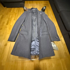 Grå ullrock från Dressmann, stl 46R - Snygg grå rock från Dressmann i ullblandning, klassisk rak modell med krage och knappar. Fodrad insida och avtagbar quiltad väst med dragkedja för extra värme. Perfekt för kyliga dagar och stilren look.