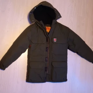 Grön dunjacka från Fjällräven XS  - Säljer en grön dunjacka från Fjällräven med huva och flera praktiska fickor. Jackan har quiltad insida, dragkedja och kardborreknäppning framtill. Perfekt för kalla dagar och har den klassiska Fjällräven-loggan på bröstet. Passar 170-177