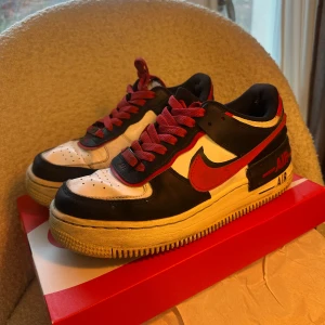 Nike Air Force 1 svart/röd/vit storlek 38 - Nike Air Force 1 sneakers i svart, rött och vitt med klassisk röd swoosh och matchande röda skosnören. Skorna har en platt sula och rund tå, tillverkade i skinn med perforerade detaljer på tån för extra ventilation. Snygg kontrast mellan färgerna och ikonisk Air-detalj på hälen.
