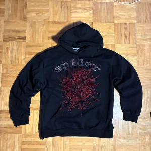Svart Spider hoodie med röd glittertryck - Svart hoodie från Spider i storlek L, tillverkad i 100% bomull. Framsidan har ett stort rött spindelnätsmotiv med glittriga detaljer och texten 'spider' i silverglitter. Tröjan har huva och långa ärmar, perfekt för dig som gillar coola och unika prints.