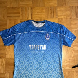 Blå fotbollströja Trapstar strl L - Snygg blå fotbollströja från Trapstar med vit logga och text på bröstet samt stort nummer 22 på ryggen. Tröjan har korta ärmar, rund hals och ett mönster i olika blå nyanser. Materialet är lätt och ventilerande mesh, perfekt för träning eller match.