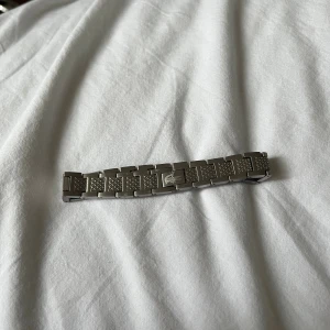 Silverfärgad länkarmband från Lacoste - Säljer ett stilrent länkarmband  från Lacoste i silverfärgat . Armbandet har rektangulära länkar med texturerade detaljer och klassisk Lacoste-logga på spännet. Perfekt för dig som vill ge din klocka en sportig och clean look. Endas en kvar skynda på 